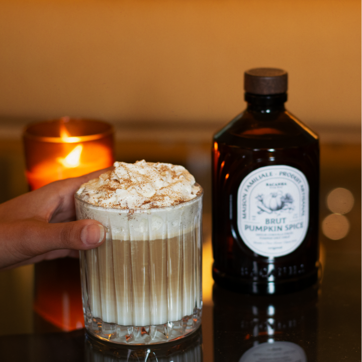 Glass of pumpkin spice latte with whipped cream next to a bottle of pumpkin spice syrup on a reflective surface.